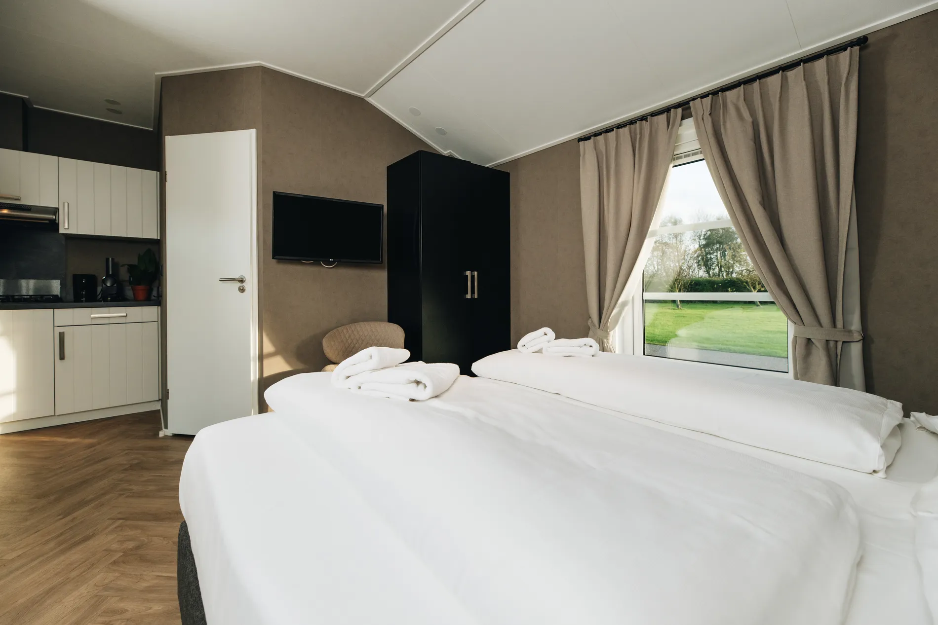 Lichte slaapkamer in een vakantiehuis met een opgemaakt tweepersoonsbed, frisse witte handdoeken, een zwarte garderobekast en zicht op de open keuken.